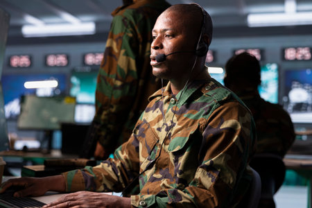 Agent in command post uses gear to identify threats from enemies. African american army operator in base of operations uses headset mic to communicate information to generals on war front lineの写真素材