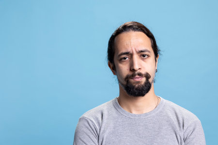 Serious person doing disappointing facial expressions on camera, pondering and posing with arms crossed against blue background. Male model with a natural authentic attitude in studio.の写真素材