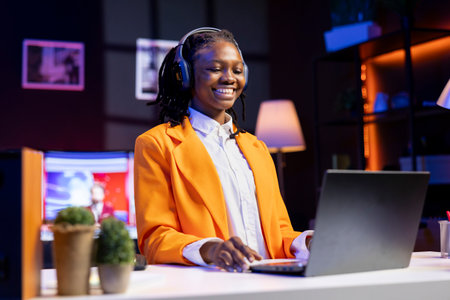 Woman sitting at desk wearing headphones laughing, watching funny TV shows on laptop, streaming content. Person entertained by TV series on chroma key notebook display, selecting episodesの写真素材