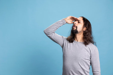 Young cool person with long hair and beard shielding his eyes to search for someone in the distance, observing and exploring against a blue background. Focused inquisitive man conveying curiosity.の写真素材