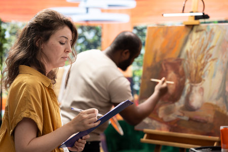 Female painter takes notes and teaches the art of painting in atelier, analyzing the creative technique of her apprentice. Artist helping man to create a nature inspired paintwork in workshop.の写真素材
