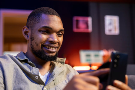 Relaxed african american guy texting his friends over social media apps, using his mobile phone to navigate on the internet. Young adult sitting in his living room and scrolling web.の写真素材