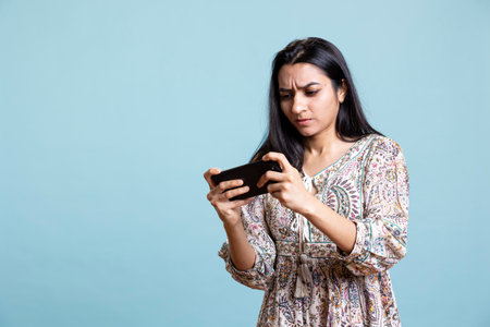 Happy indian woman entertained by videogames on smartphone, enjoying leisure time. Gamer enjoying game on phone, having fun defeating enemies, standing against studio background.の写真素材