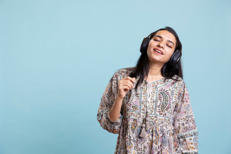 Happy woman playing electronic music and waving hand in the air, doing groovy dance moves in the studio. Cheerful confident girl wearing headphones and listening to her songs on camera.の写真素材