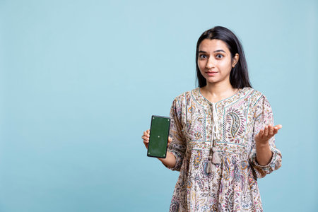 Portrait of indian woman doing marketing ad with green screen device, standing against blue background. Smiling young adult feeling positive on an advertisement in front of the camera.の写真素材