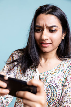 Indian girl playing entertaining mobile video games in studio, celebrating win while she stands against studio background. Young adult loving to play gaming competition.の写真素材