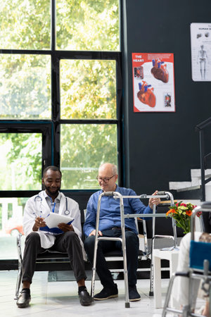 Black medic doing x ray scan interpretation with elderly man in the lobby, discussing about recovery steps and medication. Senior patient with reduced mobility uses a waling frame, evaluation.の写真素材