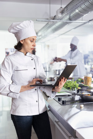 Dedicated woman chef using laptop for research and tips in cooking gourmet meals, restaurant kitchen balancing technology and tradition. Expert chopping vegetables and raw materials.の写真素材