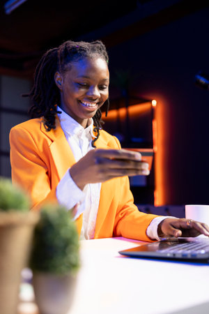 Smiling woman excited to do online shopping, typing debit card information on laptop in apartment. Cheerful person at desk using notebook, adding payment method on websiteの写真素材