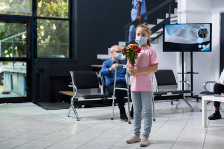 Little girl bringing flowers to grandparent in clinic lobby during medical visit, showing her support and love on the road to recovery. Safety mask regulations and hygiene policy for patient care.の写真素材