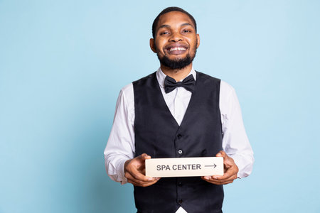 Hotel porter in suit and tie helping with directions to the spa center using a pointer, holding the wall sign to guide guests to resort amenities. Providing great concierge services.の写真素材