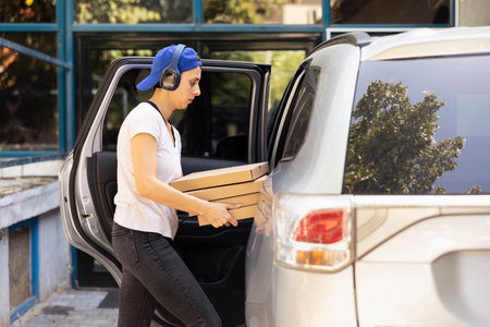 Female city courier carrying massive stack of pizza boxes delivering fast food order from a restaurant. Driving the courier service car to bring bigger meal orders, pizza delivery.の写真素材