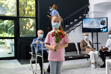 Young kid visiting elderly grandparent in medical facility waiting area, holding flowers bouquet to show love and support while wearing mask and following healthcare protection rules.の写真素材