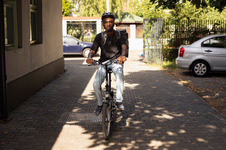 Black male courier bringing meal order to the doorstep of a client, delivering food package as an express city service. Delivery man riding the bike at locations to get food from restaurant.の写真素材