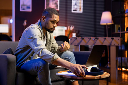 African american man grabbing his headphones to listen to music while he works, enjoys multitasking with his remote job. Person having fun with songs on headset at his modern home.の写真素材