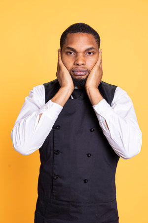 African american waiter recreates the three wise monkeys proverb, showing the symbolic metaphor by covering his ears in studio. Young restaurant employee does wisdom symbol.の写真素材