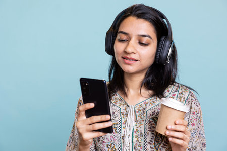 Pleased excited woman playing music on phone and drinking coffee, having fun dancing on her favorite songs against blue background. Delighted indian girl wearing headphones and doing funky moves.の写真素材