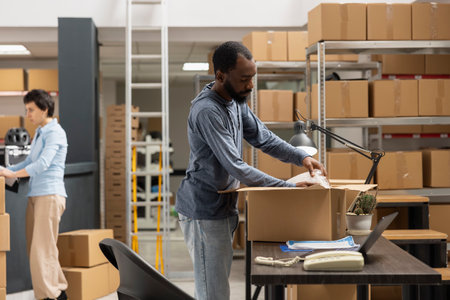 Black male employee reviewing shipment logistics for distribution hub, working in the shipping area at the depot supply room for a local small business. Managing custom orders.の写真素材