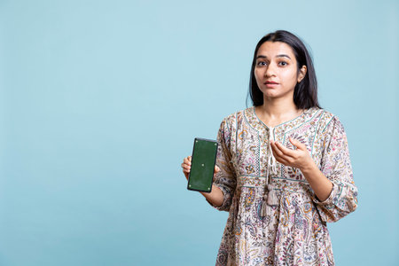Confident woman presenting green screen on mobile gadget, standing against studio background. Cheerful indian person holding copy space chroma key smartphone used for advertising brands.の写真素材