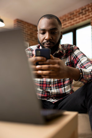 Black male remote worker taking break from laptop, using cellphone to browse social media and stay updated. Young african american professional balancing digital tasks and relaxation in home office.の写真素材