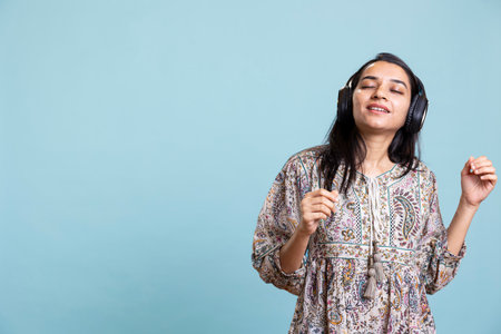 Cheerful young person listening and dancing on music with headset, singing lyrics and moving around against blue background. Happy thrilled girl wearing headphones and having fun.の写真素材
