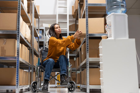 Young woman with chronic impairment working in accessible small business space, wheelchair user handles product labeling and order processing with boxes from the shelving units.の写真素材