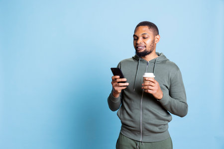 Confident relaxed person texting his friends and serving fresh cup of coffee, feeling happy and smiling at his smartphone. African american lively man messaging his pals, drink caffeine.の写真素材