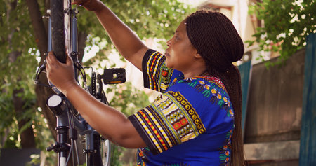 Enthusiastic athletic african american woman fastening bolts and securing wheel to bicycle frame. Dedicated female cyclist using specialized wrench to dismantle damaged bike tire outside.の写真素材