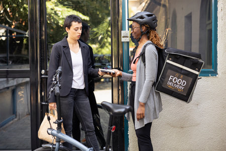 Woman completes a fast food purchase with contactless payment at the doorstep, courier holds pos terminal steady as the electronic transaction is processed. Electronic wireless purchase.の写真素材