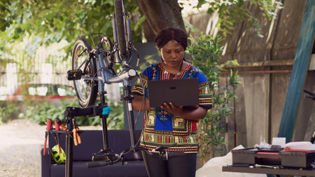Committed sports-loving woman browsing for information on modern bicycle for annual maintenance with her laptop. African american female cyclist upkeeping bike by surfing the internet.の写真素材