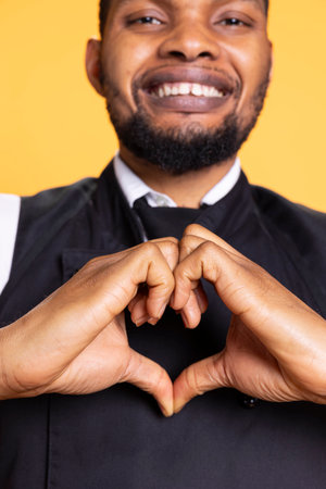 Catering staff in apron and uniform doing a heart shape sign in studio, expressing his honest feelings and true love. Restaurant worker showing a romantic gesture, sincere confident guy.の写真素材