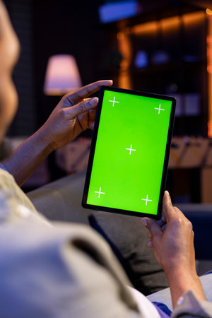Relaxed male tech user holding device with isolated mockup screen in his living room, sitting on the couch and teleworking. African american happy guy browses the internet, outsourcing.の写真素材