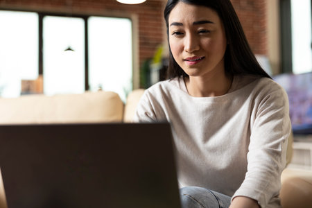 Closeup portrait of remote female manager checking emails and replying to messages on laptop. Self employed asian woman seated on sofa, studying digital reports for business growth.の写真素材
