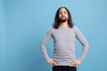 Young adult in casual wear standing with his arms crossed on camera, posing with confidence against a blue background. Man with a beard and long hair, calm natural attitude.の写真素材