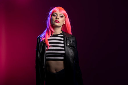 Confident female model throws bold attitude with intense eye contact and edgy styling. Pretty woman with pink hair and striped top pop posing under creative blue and magenta studio lights.の写真素材