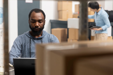 African american man overseeing product shipment and order logistics, working in small scale factory operations with taped boxes stacked on shelves. Handling goods and processing orders.の写真素材