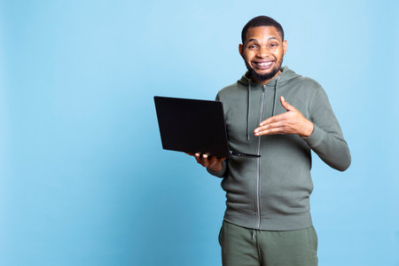 Carefree happy african american guy working on his laptop in studio, wearing a grey hoodie and composing emails to solve his tasks and paperwork. Young adult multitasking, browses websites.の写真素材
