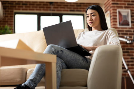 Serious female manager working remotely in brick wall apartment, typing on laptop and preparing business reports. Self employed asian woman showing productivity at home, reviewing company proposalsの写真素材