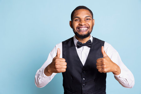 Cheerful positive hotel concierge giving a thumbs up sign on camera, presenting his approval and trust. Satisfied happy personnel in a tuxedo showing an okay like gesture, good vibes.の写真素材
