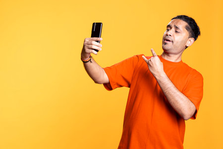 Indian young person using his mobile phone to take selfies from all angles. Male social media user taking pictures using his smartphone camera, smiling happily against studio background.の写真素材