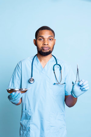 Portrait of african american medic holding surgical equipment, preparing to do medical procedure or operating with sterile tools in studio. Hospital staff member using tweezers.の写真素材
