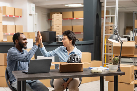 Diverse cheerful employees celebrating successful order processing task with a high five inside a small business fulfillment center, surrounded by packages and shelves in a local depot.の写真素材