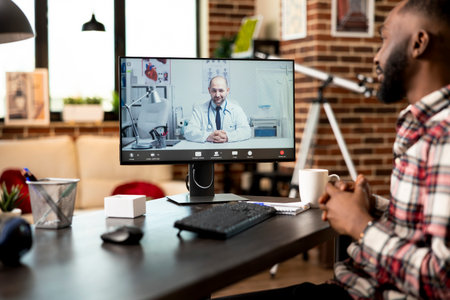 Black male employee seated at desk working remotely, describes symptoms during virtual consultation with caucasian doctor. Professionals discuss telemedicine project from home office via video call.の写真素材