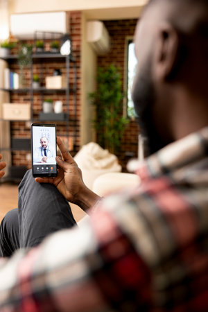 Caucasian doctor seen on device screen, conducting online health consultation with black male patient. Rearview of businessman holding cellphone, giving symptoms on video call with general practitionerの写真素材