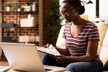Determined female entrepreneur carefully reviewing printed documents during remote work session. Focused black woman flips through clipboard, preparing notes for upcoming project presentation.の写真素材