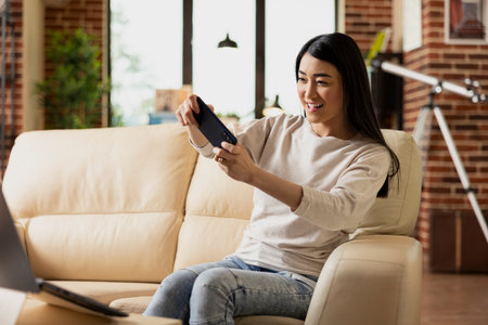 Confident self employed woman relaxes on sofa while playing video game on smartphone. Asian female entrepreneur with mobile device, enjoying fun break from work in cozy brick wall living room.の写真素材