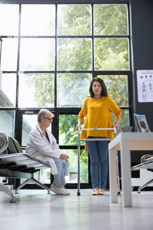 Asian woman using walking frame during rehabilitation healing process, receiving physical support from medical specialist at a health clinic. Moving around for recovery journey.の写真素材