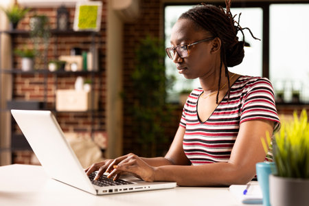 Self employed black woman working on laptop, updating her personal business platform in home office. Focused female manager reviewing digital financial files and managing project tasks.の写真素材