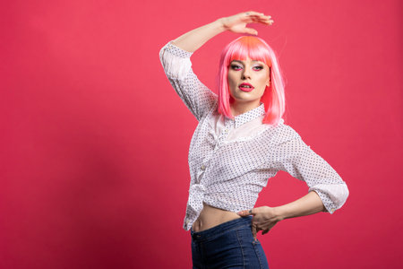 Gorgeous caucasian model with pink hair confidently striking a pose with one hand over head and other on waist. Beautiful young woman standing against isolated background, exuding self assurance.の写真素材