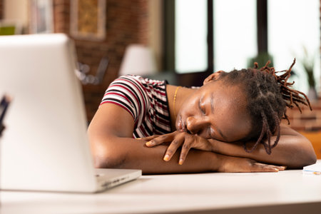 Black female entrepreneur lies on desk in home office, taking quick nap after demanding morning of...
